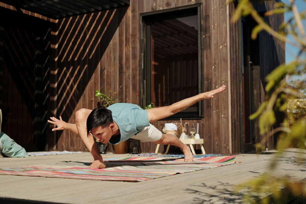 A man performs an advanced yoga pose on a colorful mat outside on a sunny veranda.